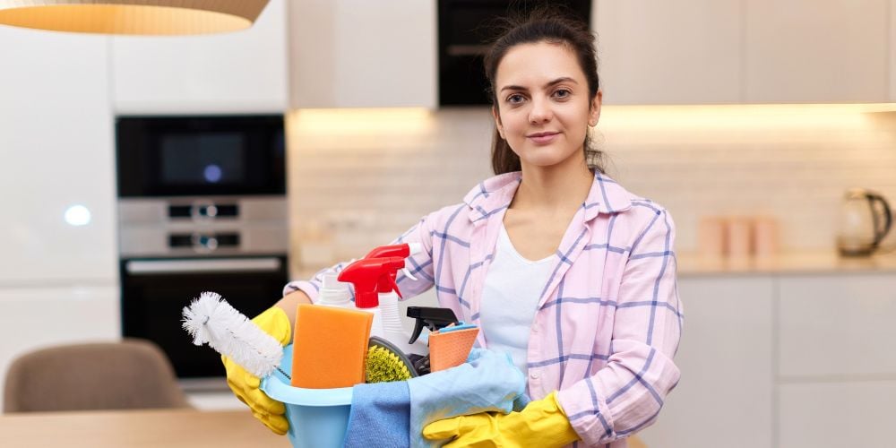 Professional house cleaner wearing yellow rubber gloves holding a basin filled with cleaning supplies in a modern kitchen.